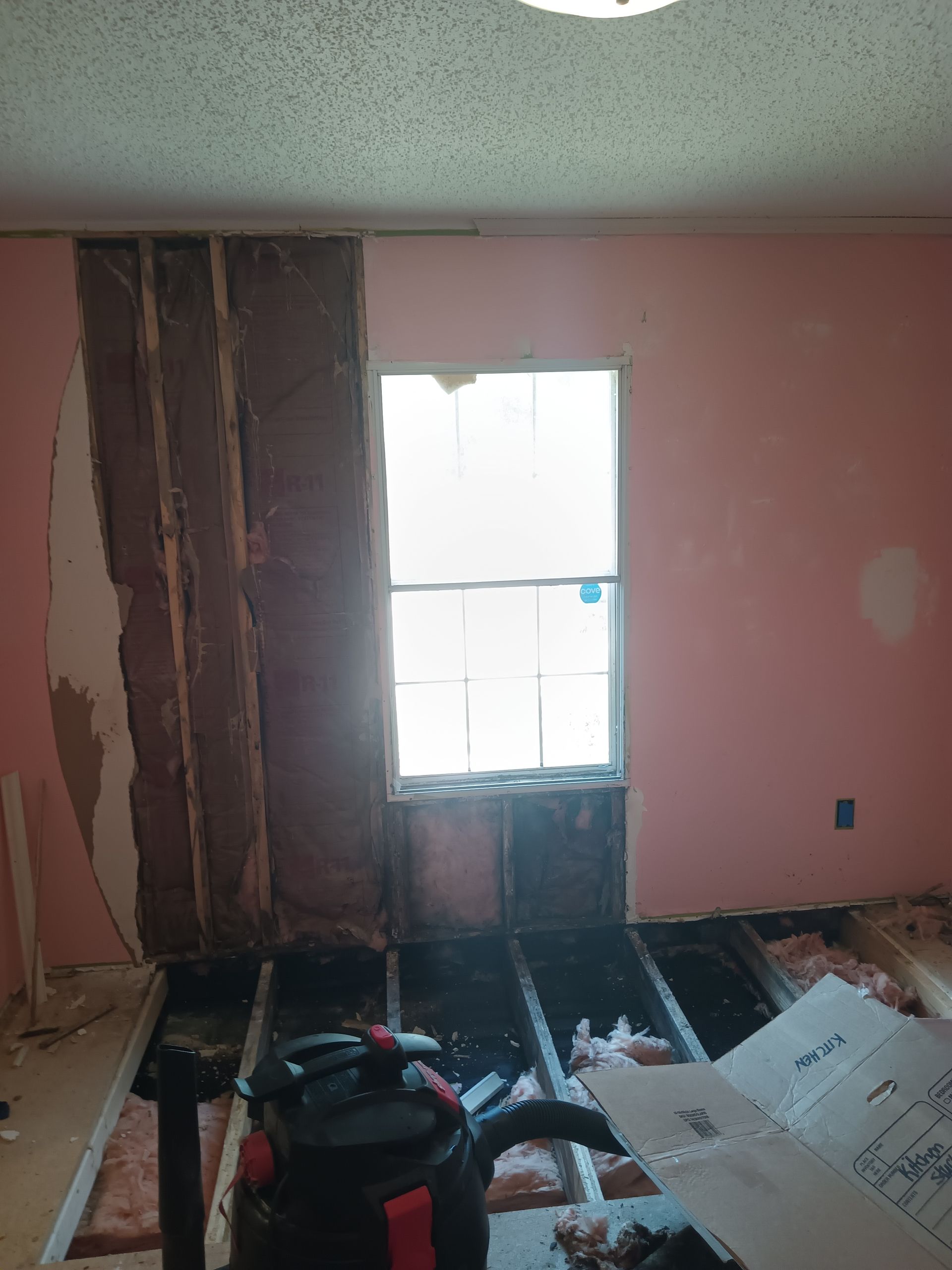 A room under renovation with exposed wall studs, insulation, and removed flooring beneath a window on a pink wall.