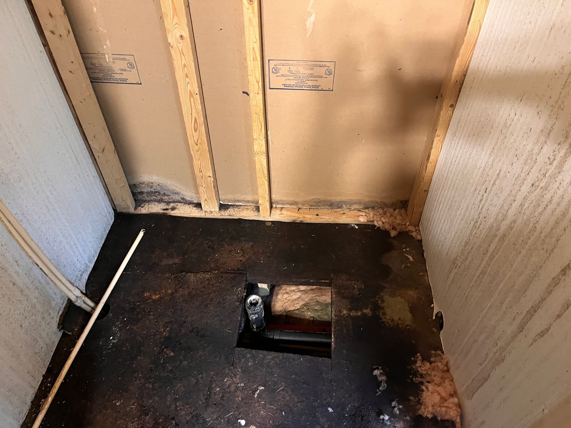 An unfinished bathroom area featuring exposed wall studs, dark subflooring, and an open hole around a plumbing pipe.