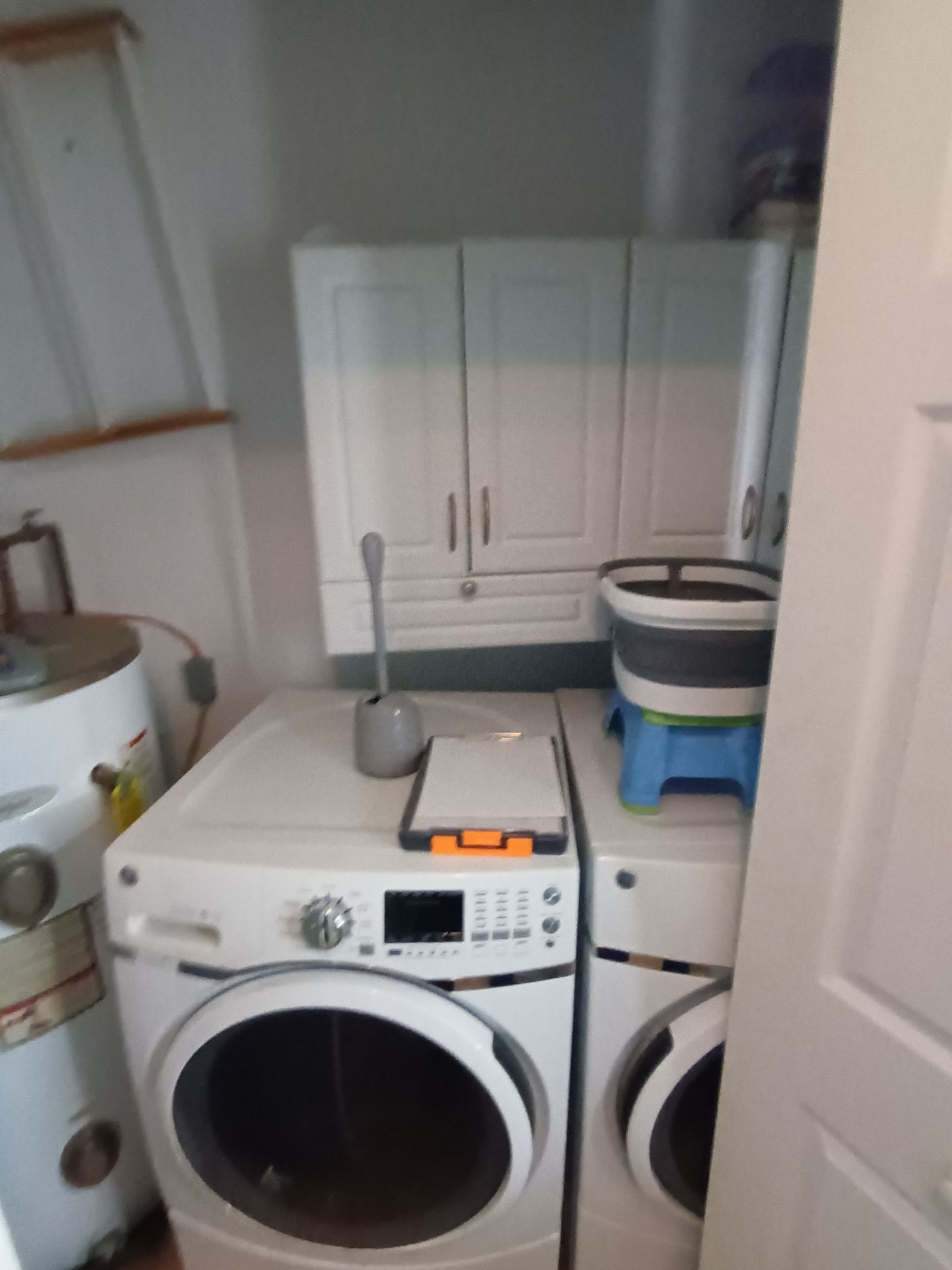A laundry room with a white washer and dryer, white wall cabinets, a water heater, and a blue collapsible laundry basket.