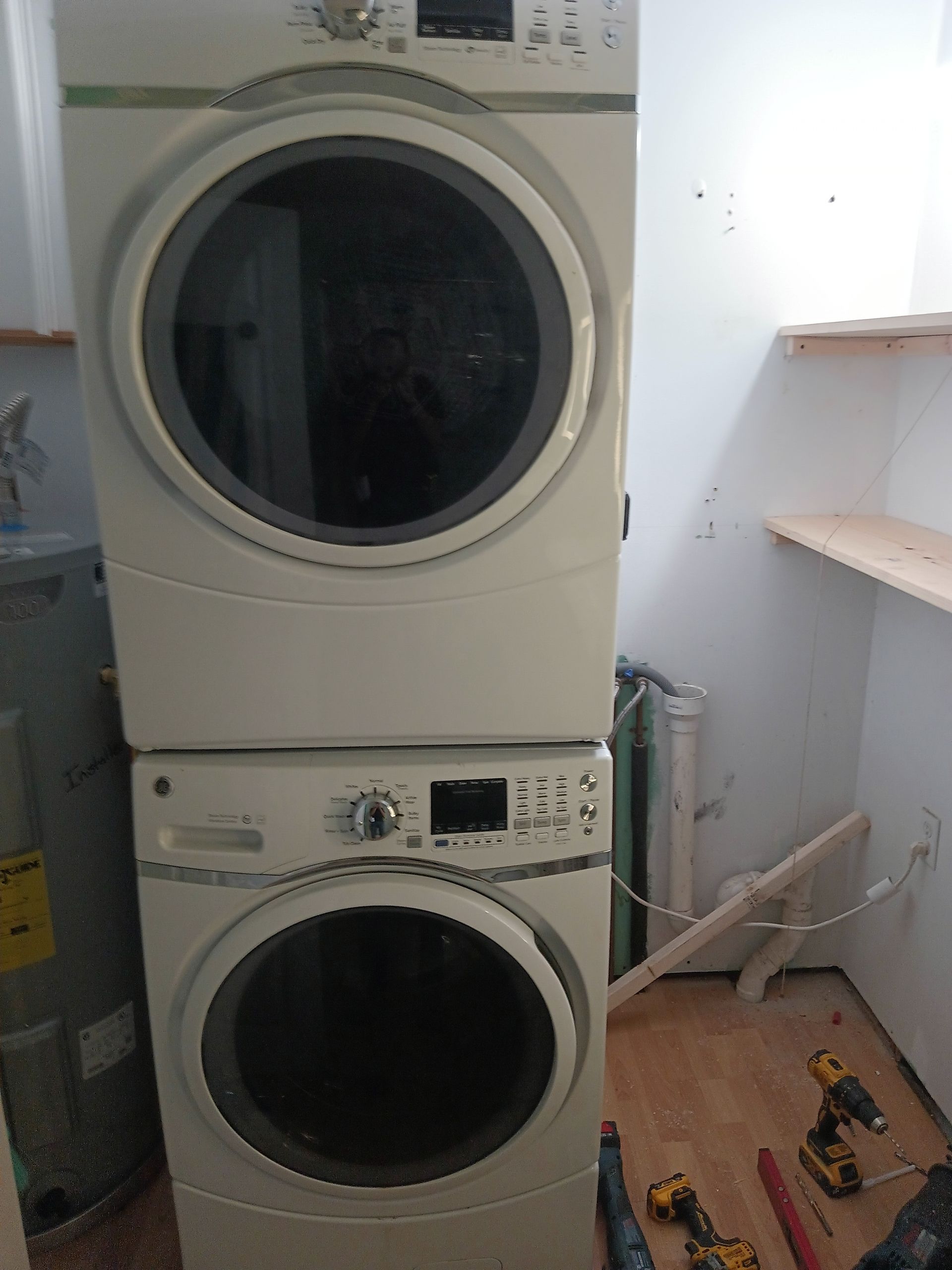 A white stacked washer and dryer unit in a room under construction with tools and shelving visible on the floor and wall.