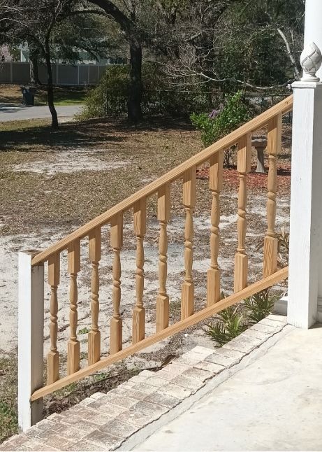 A wooden staircase railing with decorative spindles installed at an angle on a concrete porch against a grassy yard.