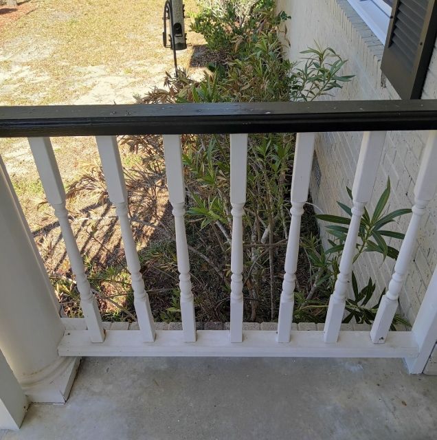 A porch railing with white wooden balusters and a dark handrail, overlooking a patch of green shrubs and sandy ground.
