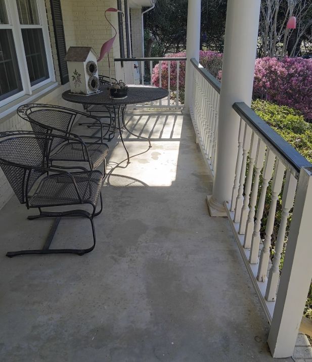A metal patio set with two chairs and a table sits on a concrete porch next to a white railing and blooming azaleas.