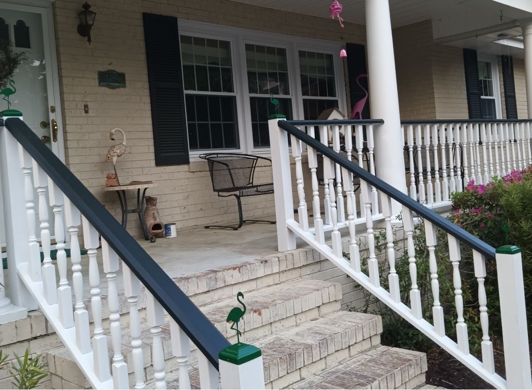 Front porch with white railings, dark handrails, brick stairs, and black window shutters on a light-colored brick house.