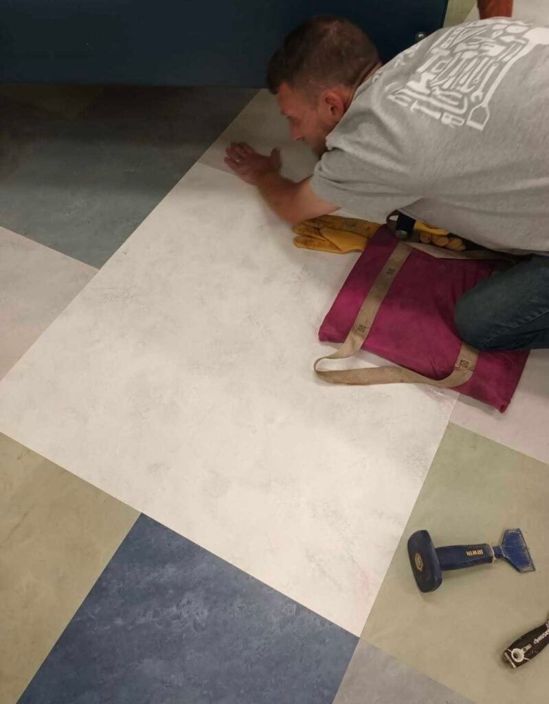 A person kneeling on a floor installs large, multicolored square tiles, using tools nearby to align the material.