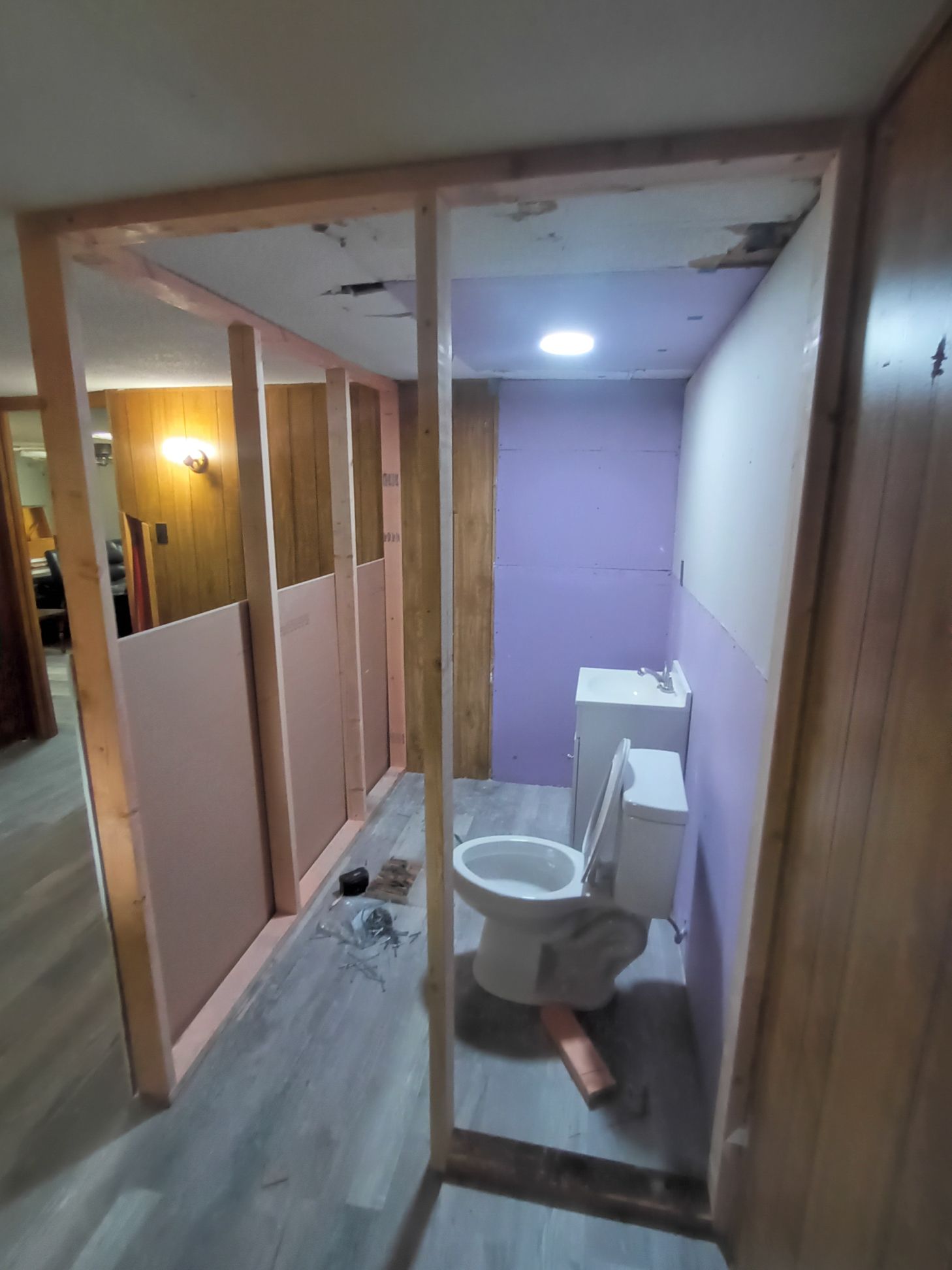 A wooden frame partitions a room containing a toilet and purple drywall in a home under construction with wood paneling.