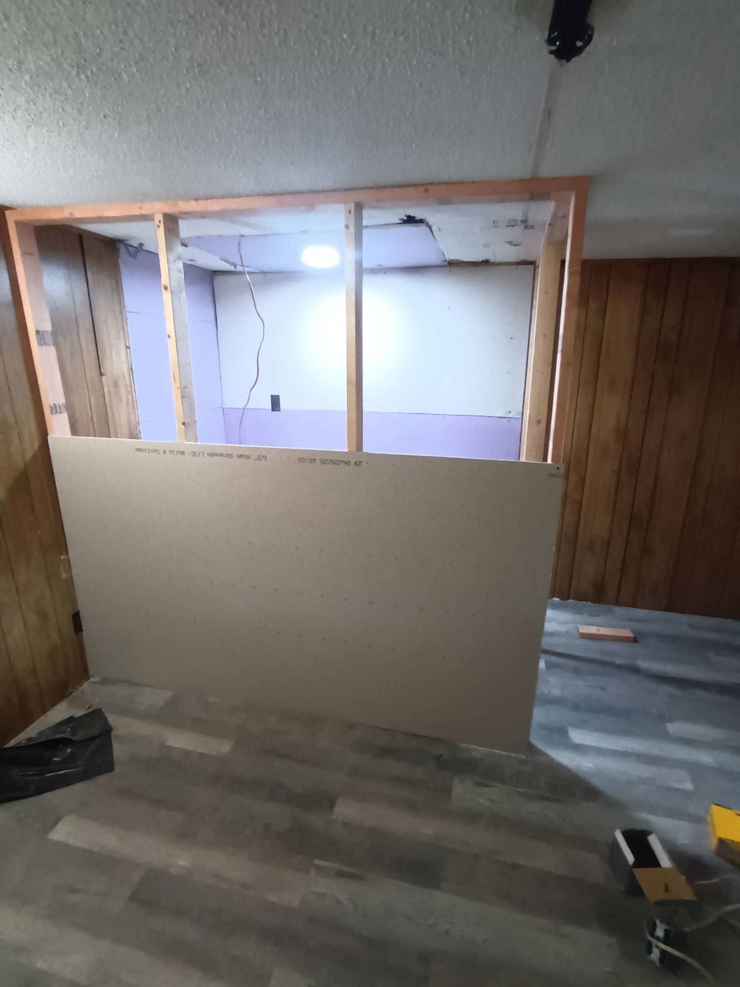 A wooden partition wall frame under construction, featuring a partially installed drywall panel in a room with wood paneling.
