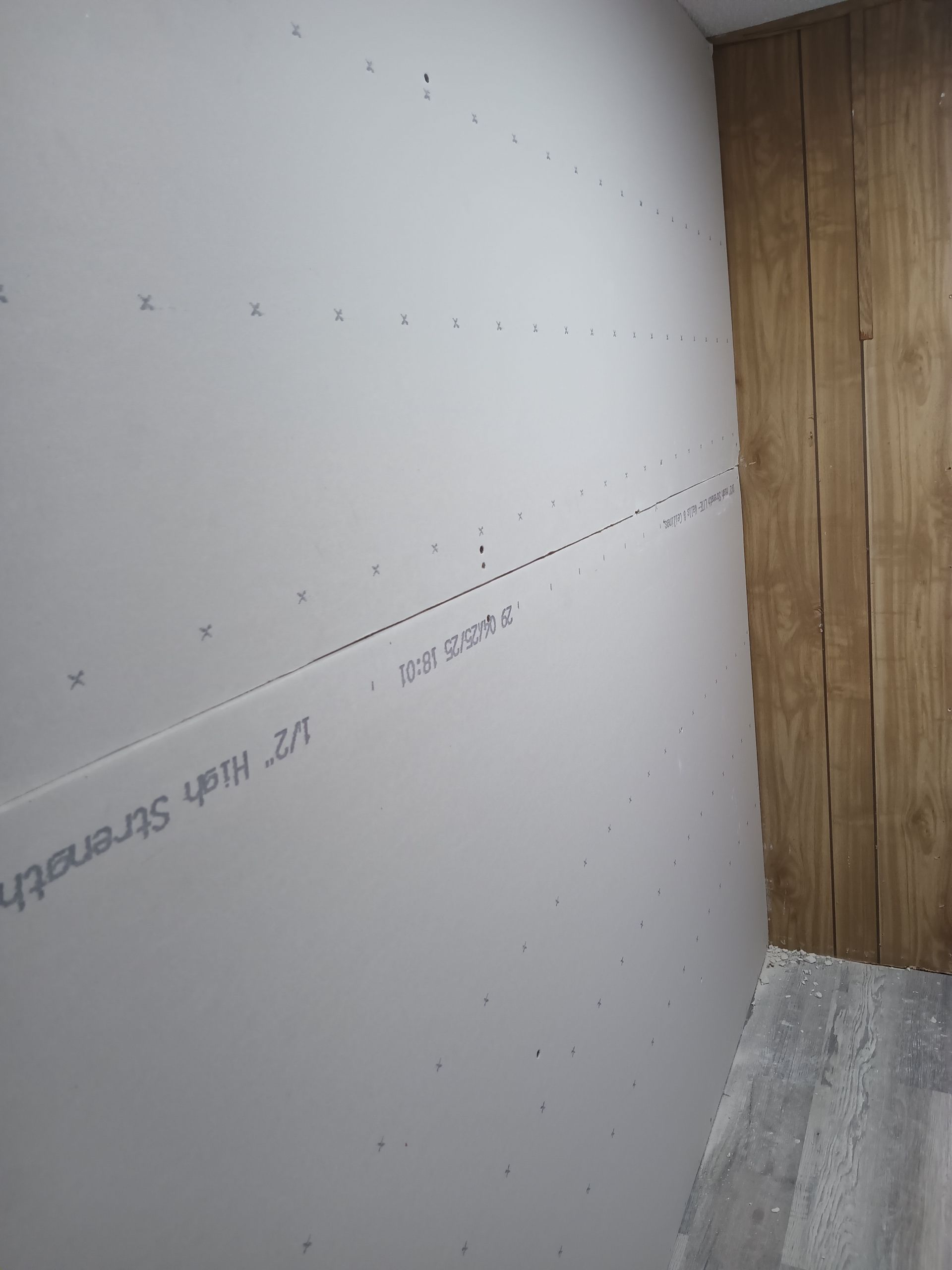 A newly installed drywall panel featuring visible screw patterns next to a wood-paneled wall and light-colored flooring.