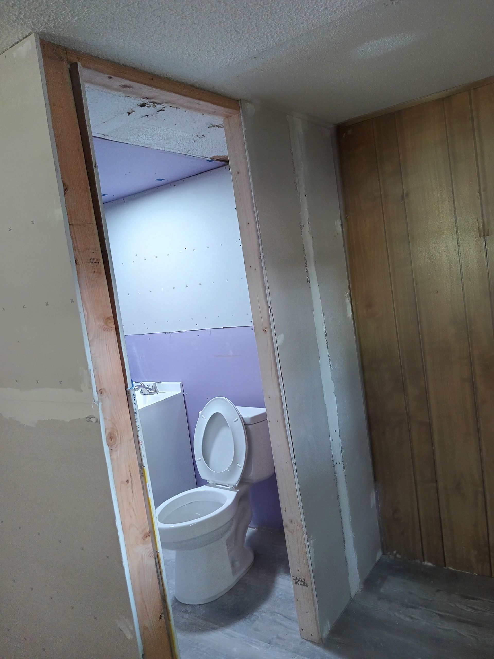 A doorway opening reveals a toilet in a room with light purple walls and unfinished drywall.