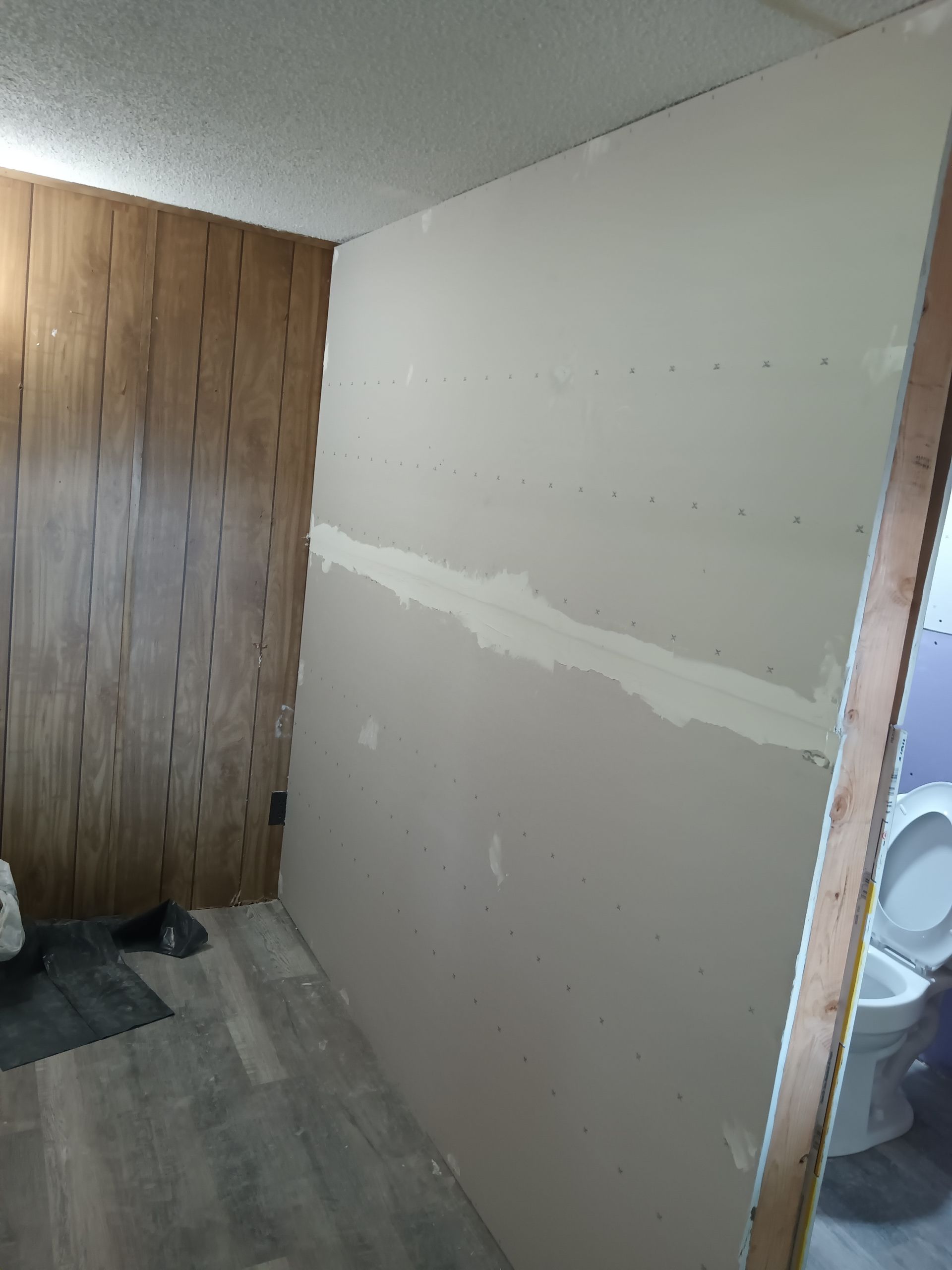A partially renovated interior wall with drywall seams, standing next to wood-paneled walls and a doorway to a bathroom.