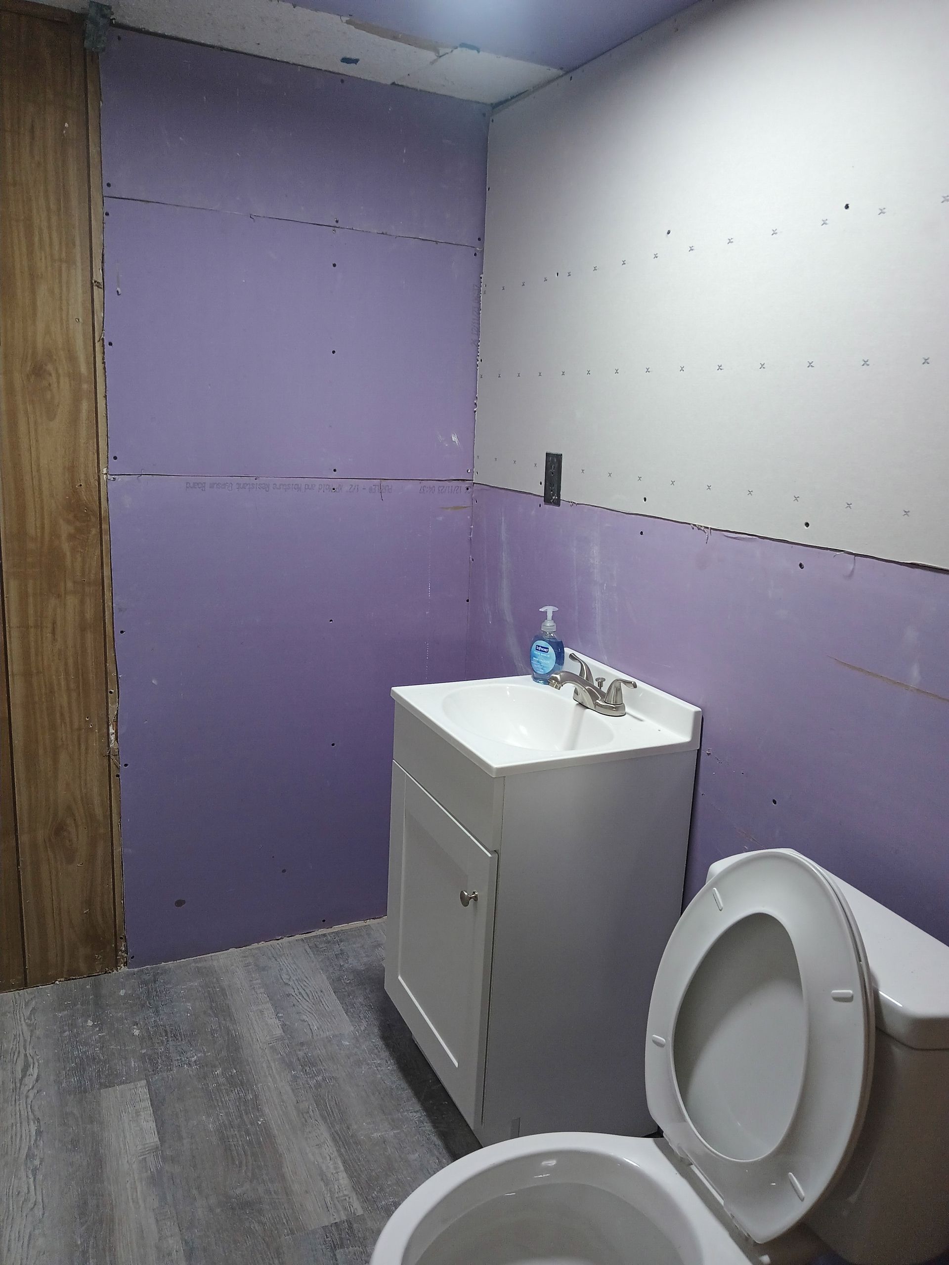 A bathroom under renovation with purple drywall, a white vanity sink, a toilet, and gray wood-look flooring.