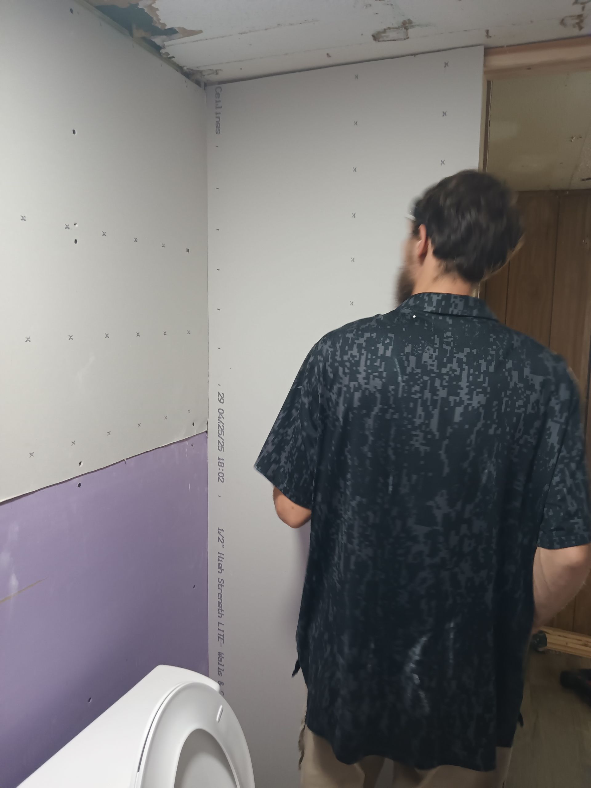 A person stands in a room undergoing renovation, facing a wall newly installed with grey and purple drywall panels.