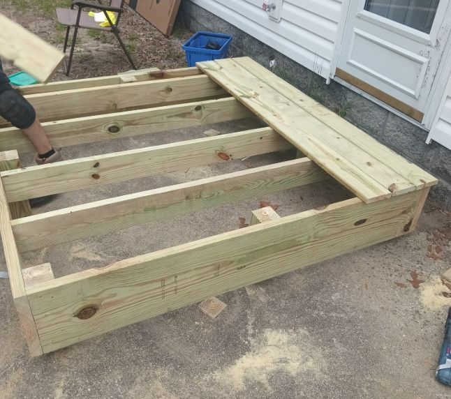 A person building a wooden deck frame outdoors on a concrete patio next to a house.