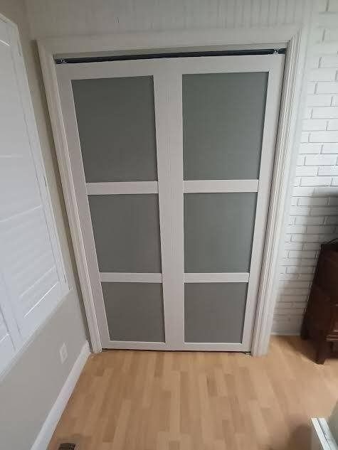 A pair of white bi-fold closet doors with three grey panels on each door, set against a white brick wall and wood flooring.