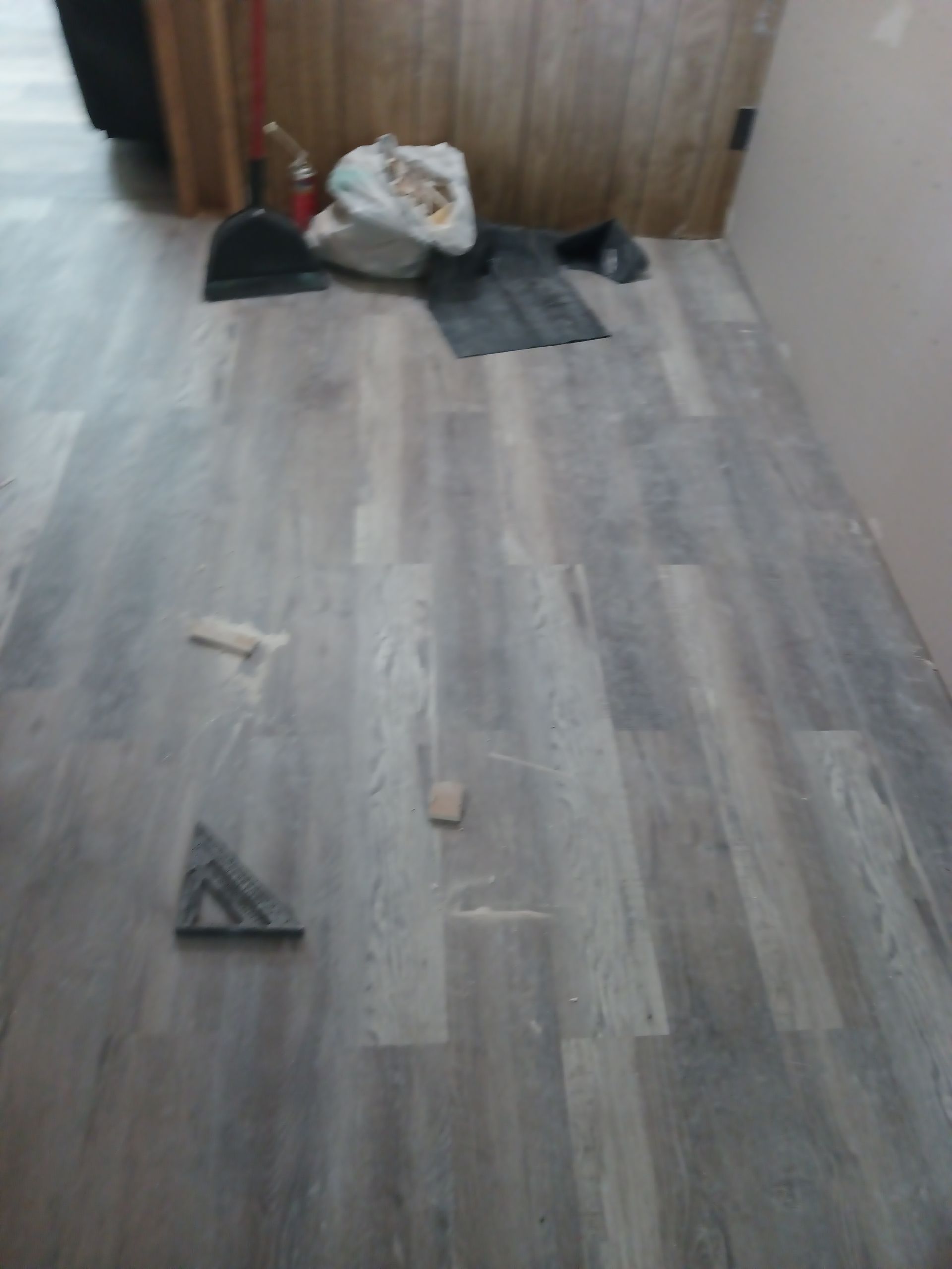 A partially installed gray wood-look laminate floor in a room, with a dustpan, tool, and scrap pieces scattered about.