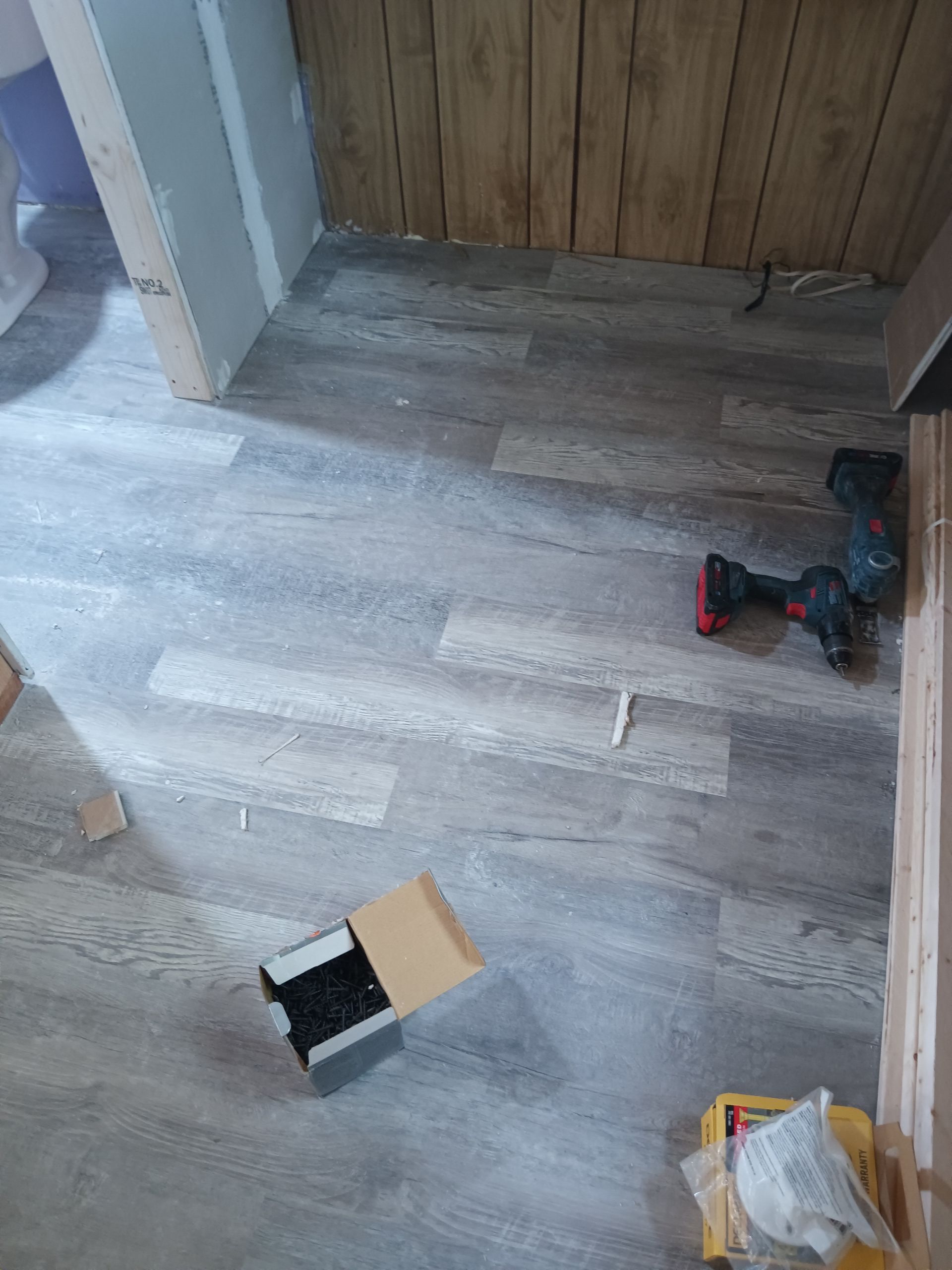Gray wood-look flooring installed in a room with a power tool, a box of screws, and a wooden paneled wall.