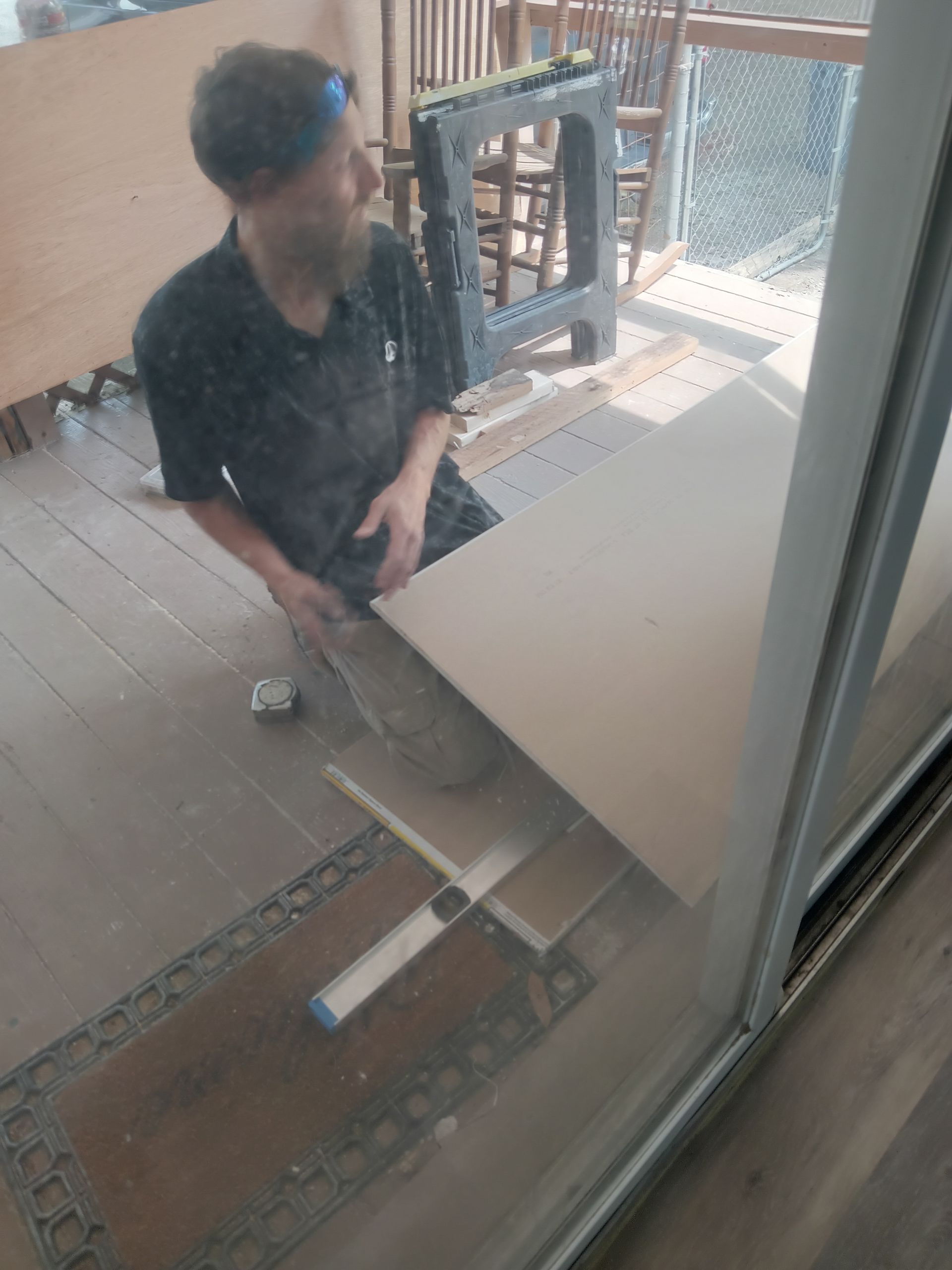 A person kneeling on a deck, using a measuring tool on a wooden board, with a black sawhorse nearby.