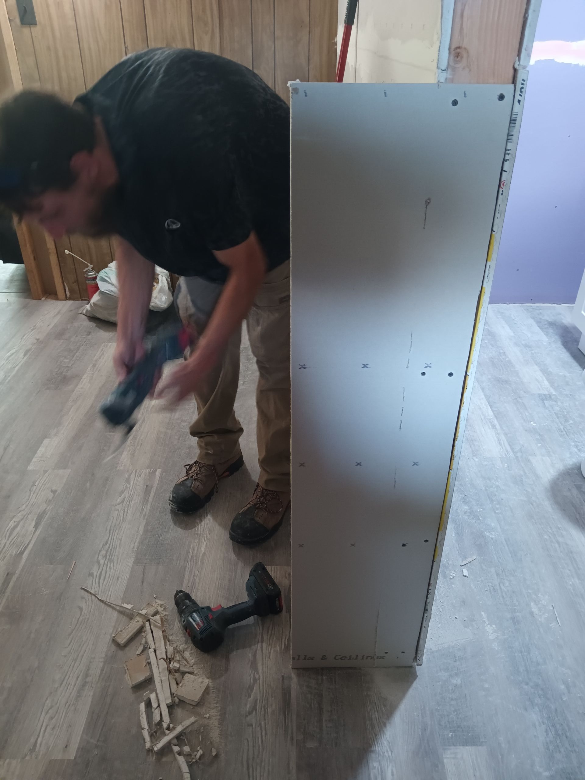 A person wearing a black shirt and tan pants uses a power tool on a wall panel in a room with wood-patterned flooring.