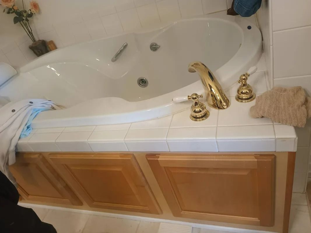 A white jetted bathtub with gold fixtures, set in a white tiled deck with wooden cabinets underneath.