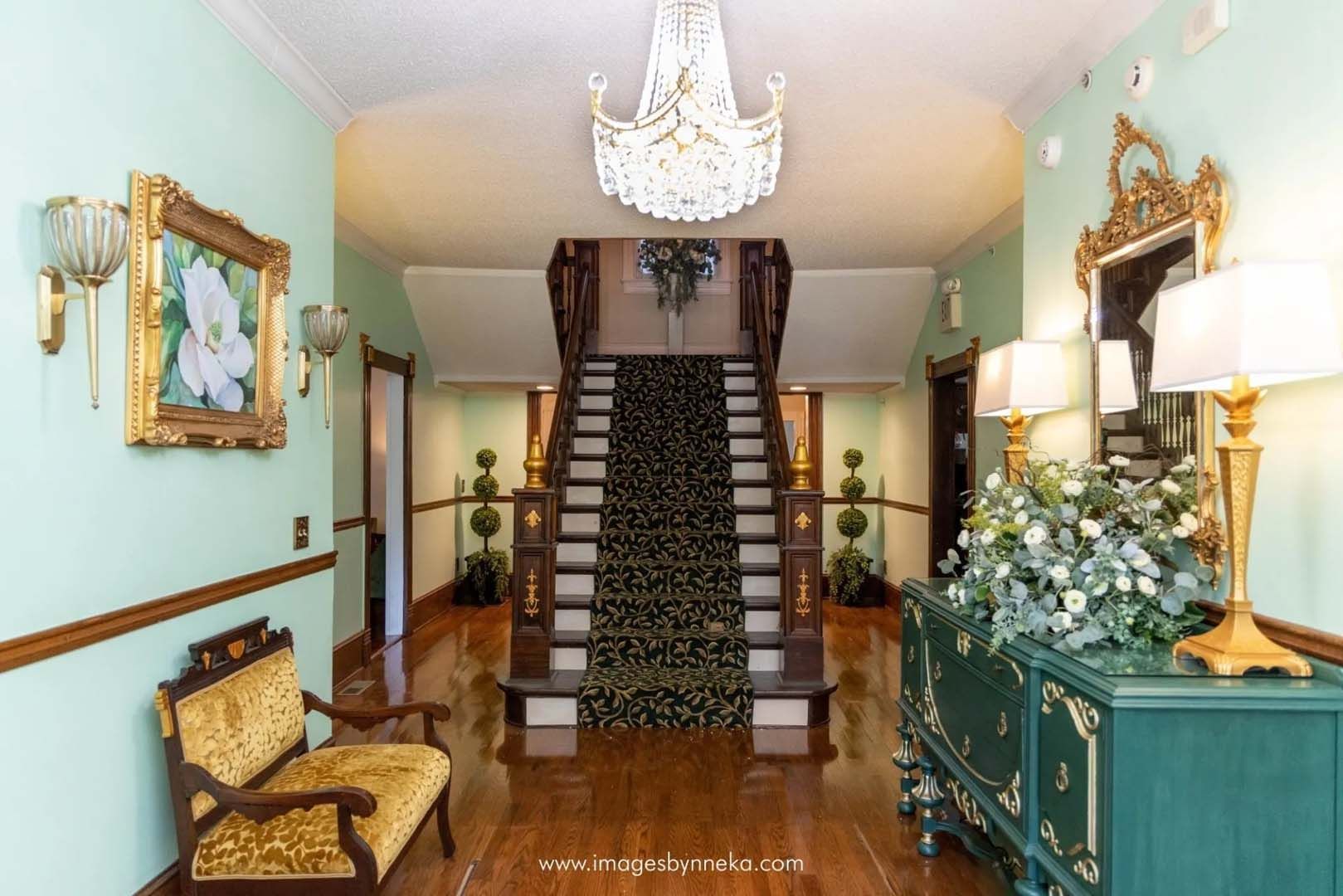 Elegant mansion foyer with grand staircase, teal walls, crystal chandelier, gold accents, and a floral console.