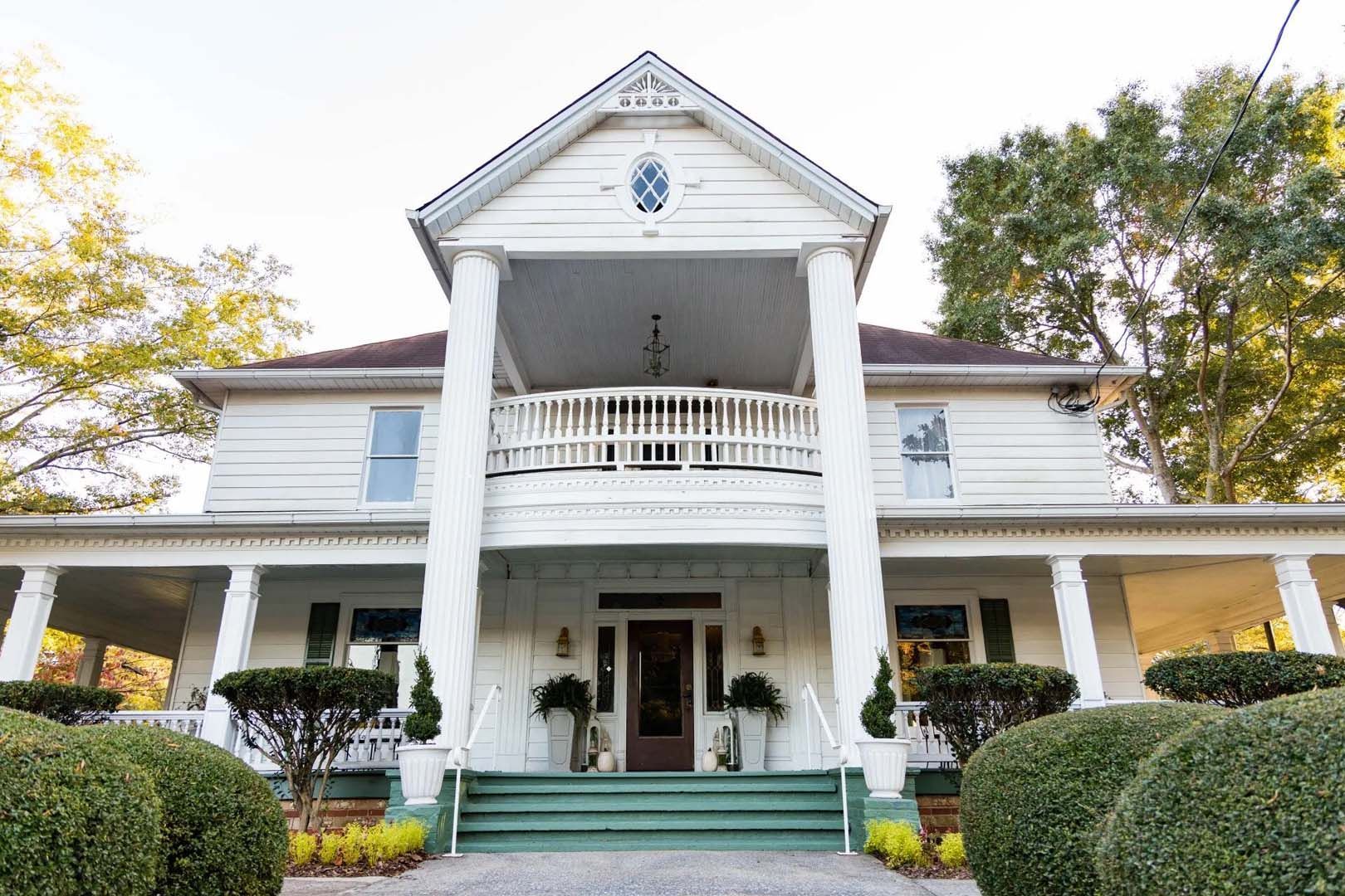 White two-story house with front porch, columns, and a balcony. Green steps lead to a dark brown door.