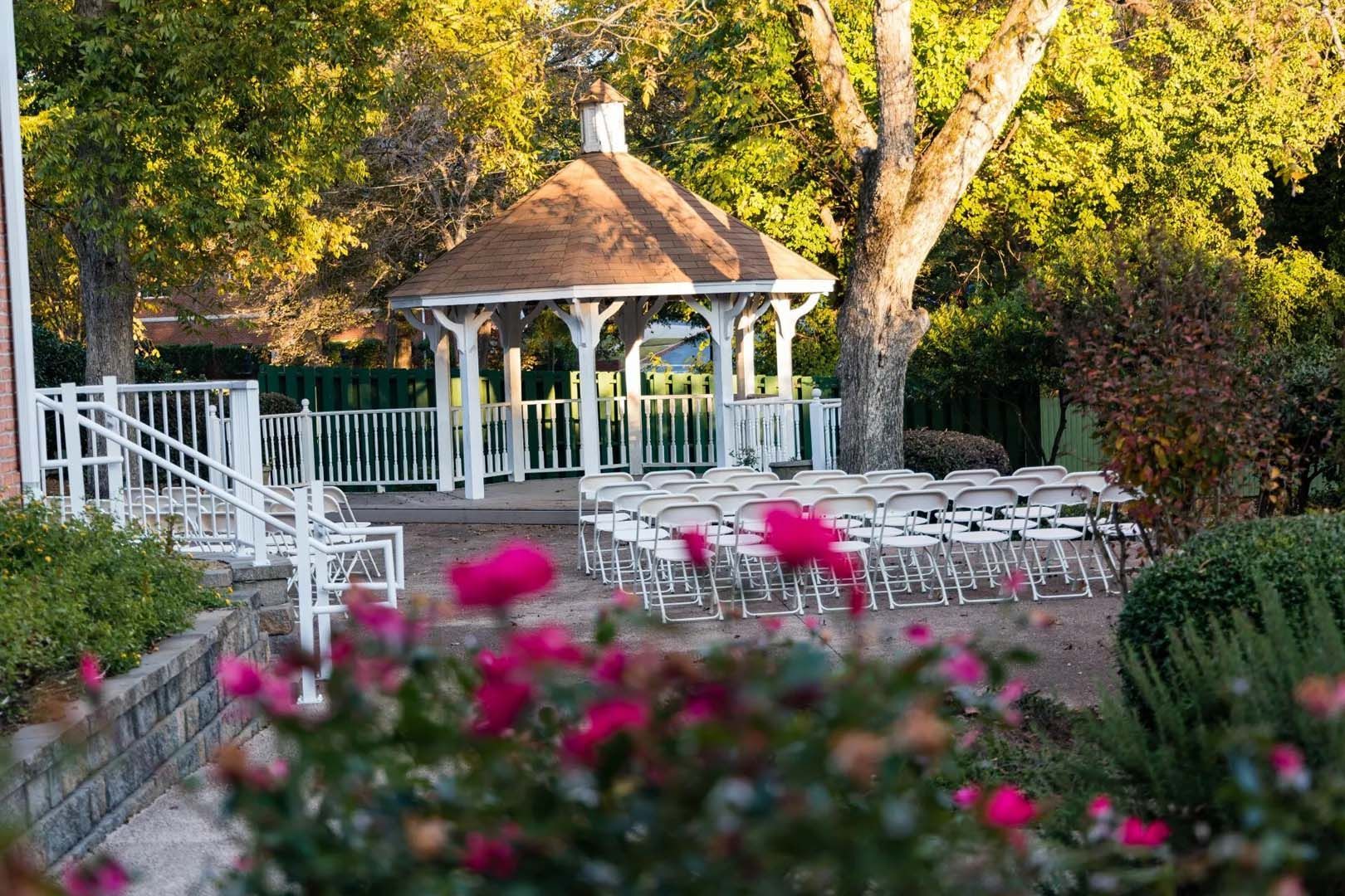 White gazebo with chairs set up for an event in a garden with pink flowers.