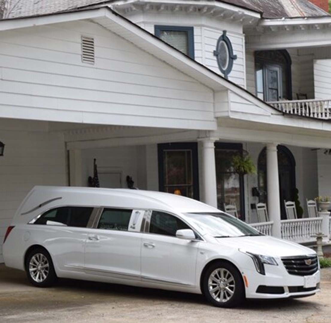White hearse parked in front of a white house with columns, likely at a funeral home.