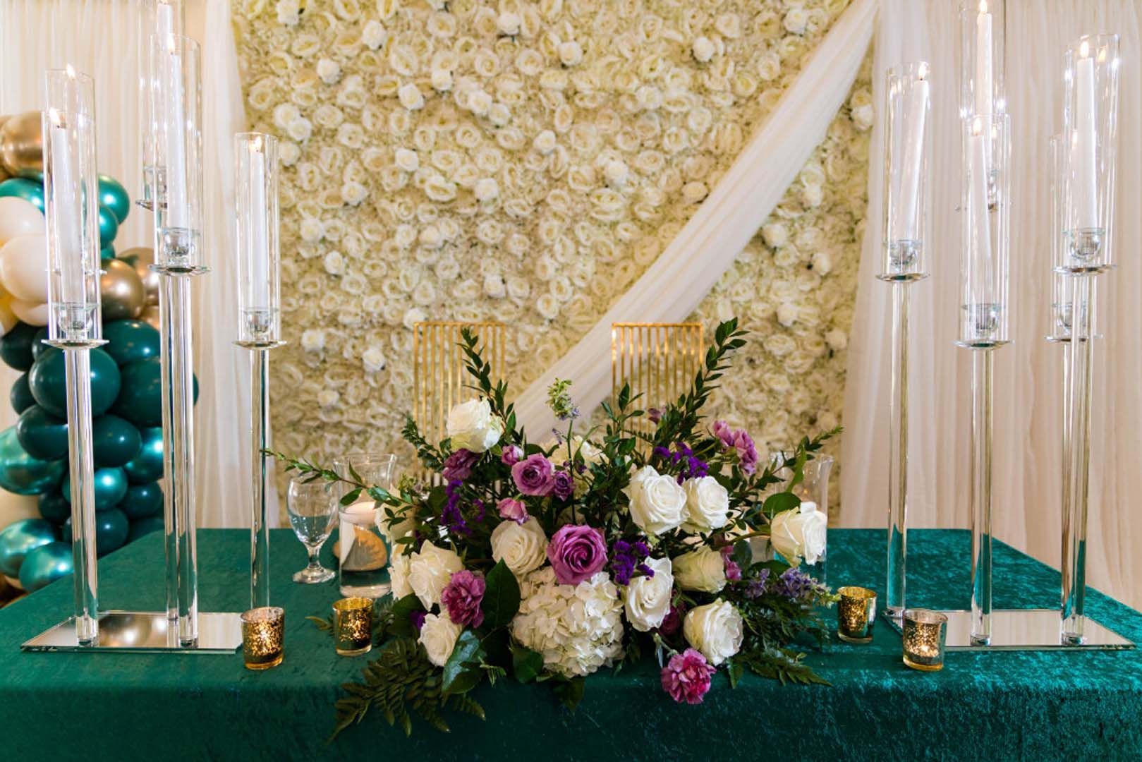 Wedding reception table with floral arrangement, candles, and teal tablecloth. White flower wall in the background.