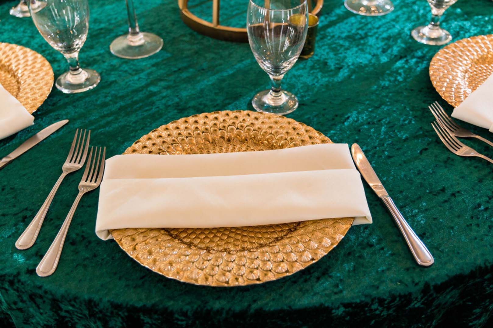Formal table setting with gold charger plate, folded napkin, and silverware on a teal tablecloth.