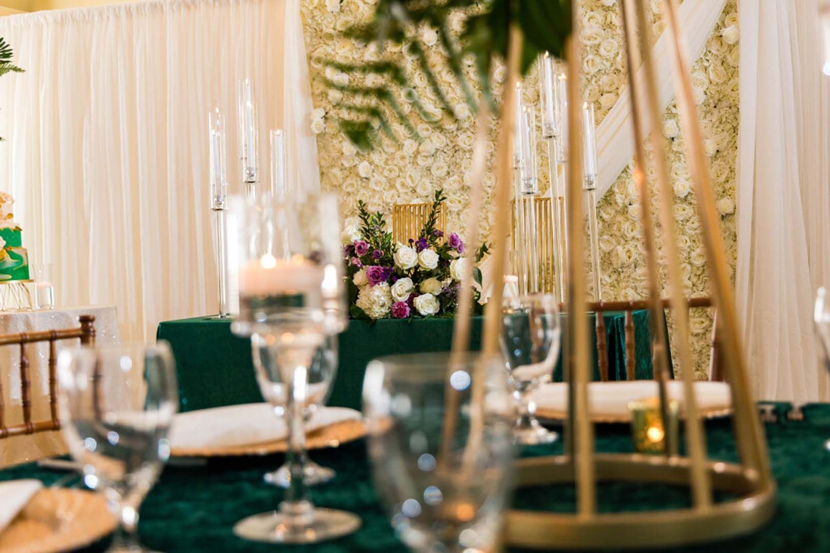 Wedding reception table with floral centerpiece, candles, glasses, emerald green tablecloth, and gold accents.