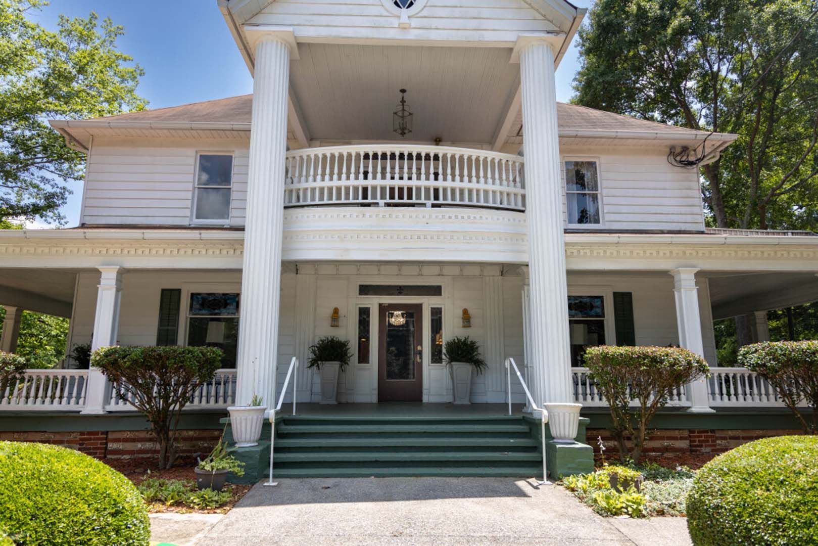 White two-story house with a porch and columns, green steps, and trimmed bushes in front.