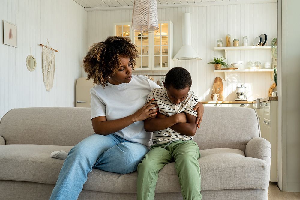 Woman comforts child on couch; both are in a light-filled living room.