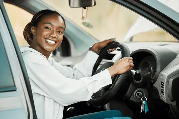 Woman smiling, sitting in a car, hands on steering wheel. Sunlight illuminates the car interior.