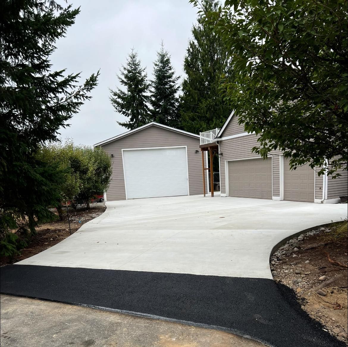 Concrete driveway leading to a house with a two-car garage. Asphalt edge, trees, and cloudy sky visible.