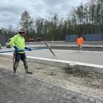 Construction worker smoothing concrete with a long tool; another person in the background. Outdoors.