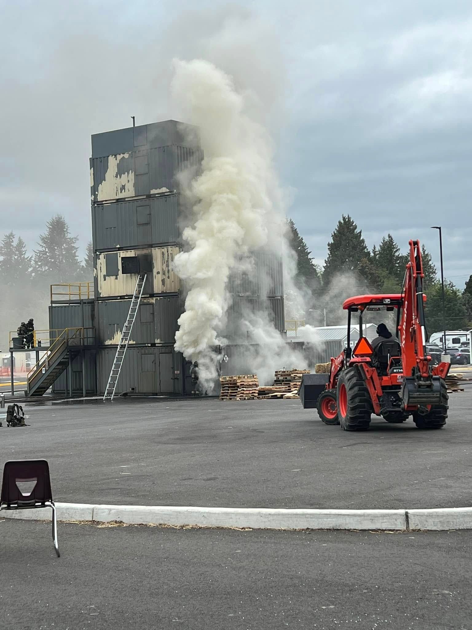 Fire training tower with smoke billowing, tractor near.