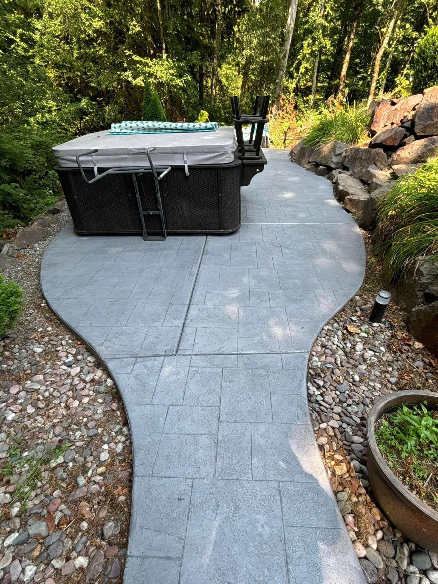 Hot tub on a patterned concrete patio with a curved pathway, surrounded by landscaping and rocks.