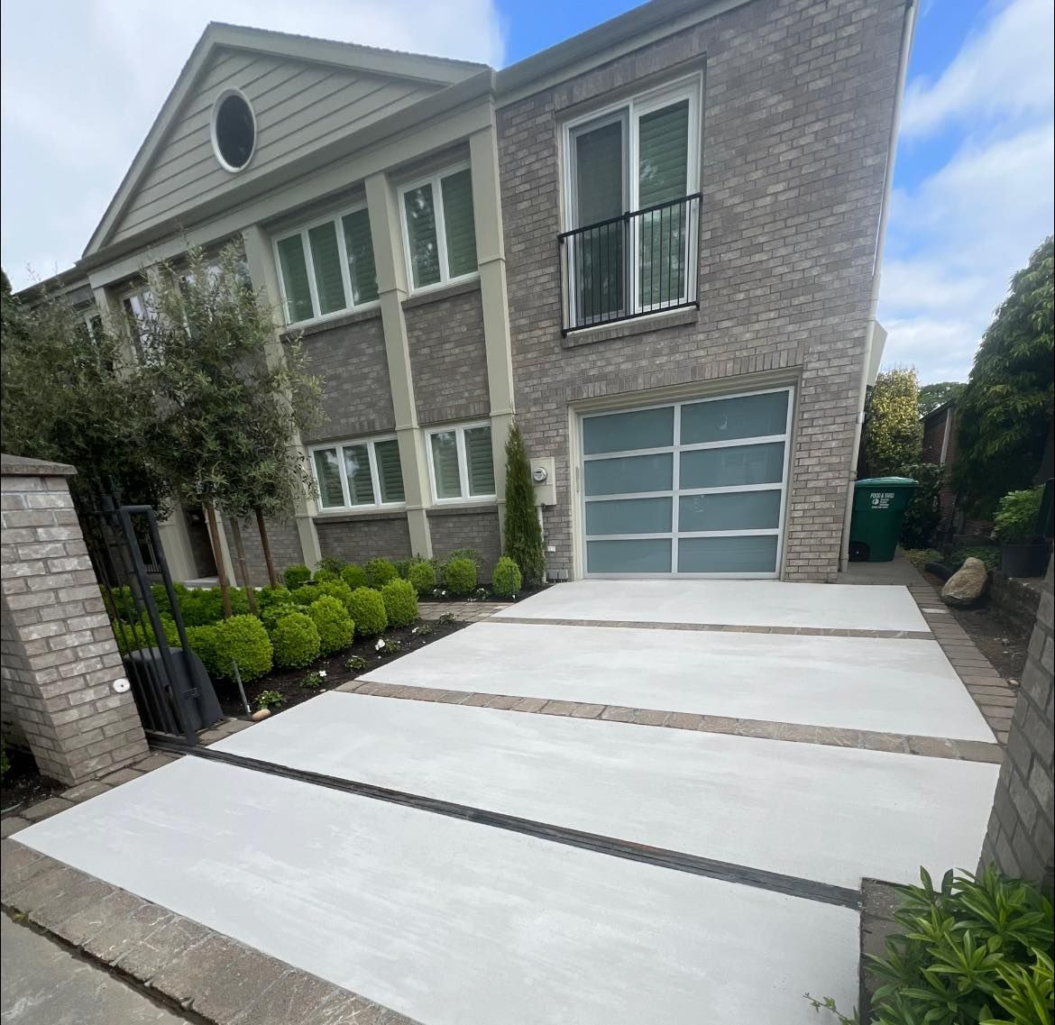 Modern gray brick house with concrete driveway, garage, and landscaping under a cloudy sky.