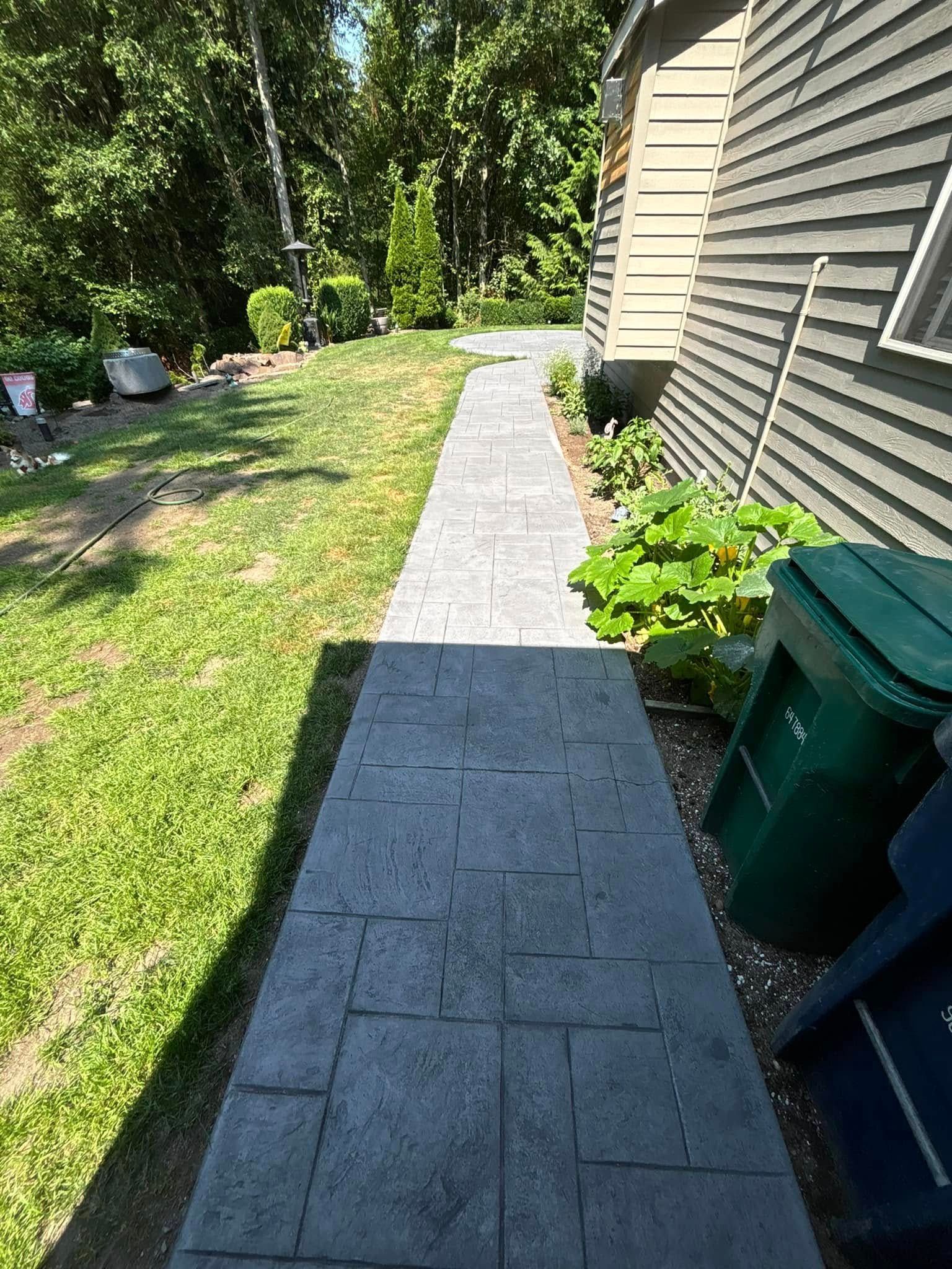 A gray stone pathway leads alongside a house with green siding and trash cans on a sunny day.