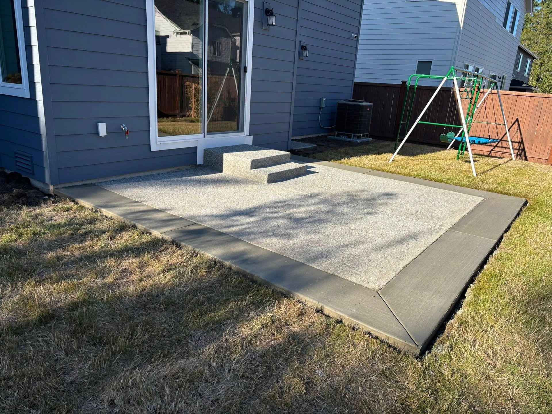 Concrete patio with border next to a house with sliding glass doors; a swing set is in the yard.