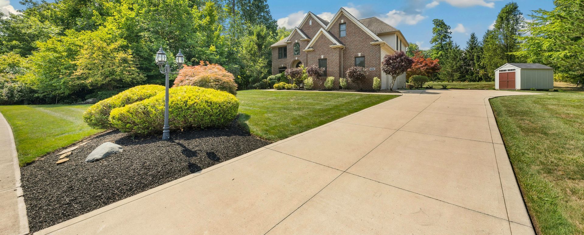 A long concrete driveway leading to a brick house with lush green lawn, trees, and shed on a sunny day.