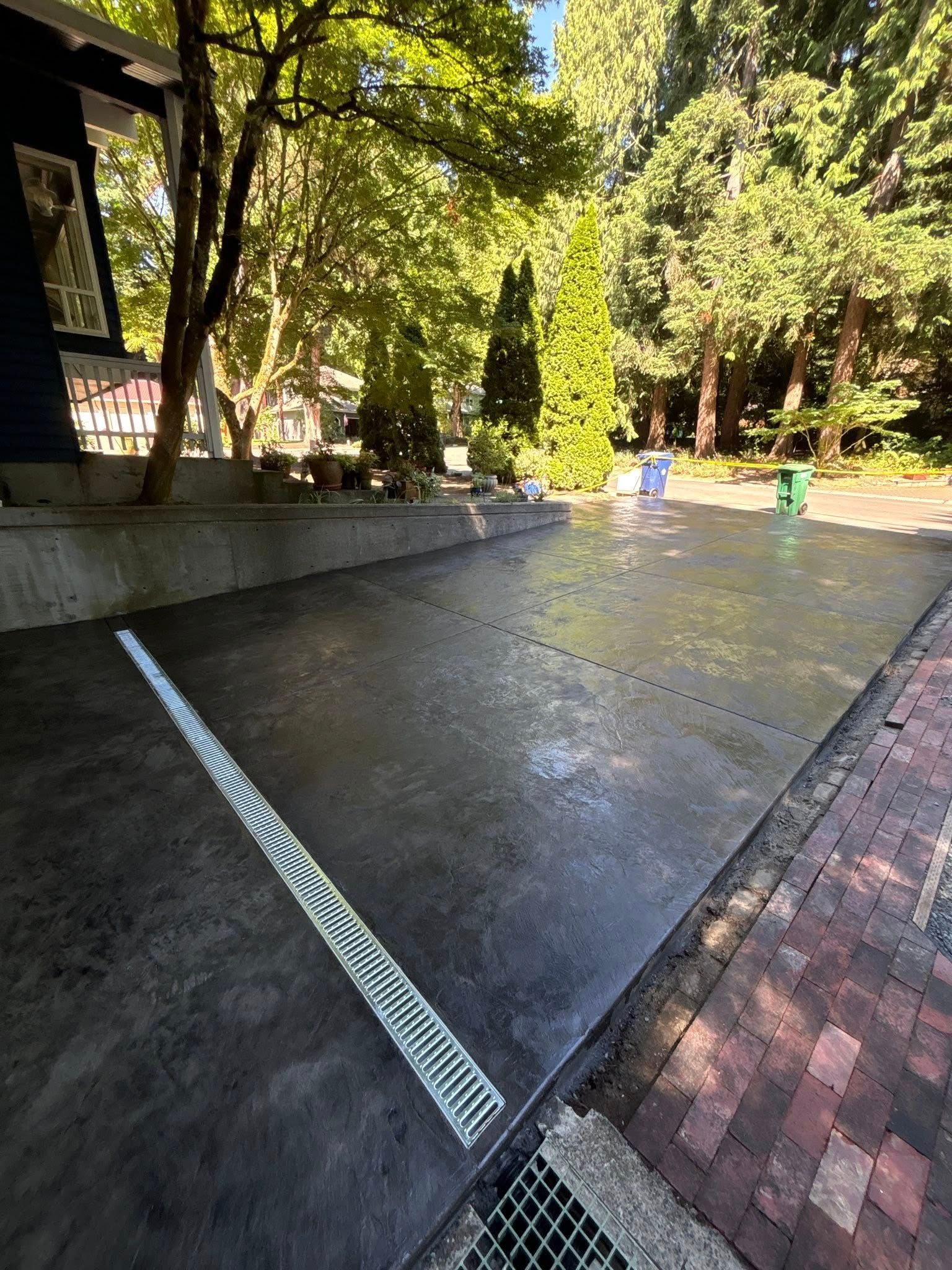 Concrete driveway with a linear drain, next to a brick path and building. Trees in the background.