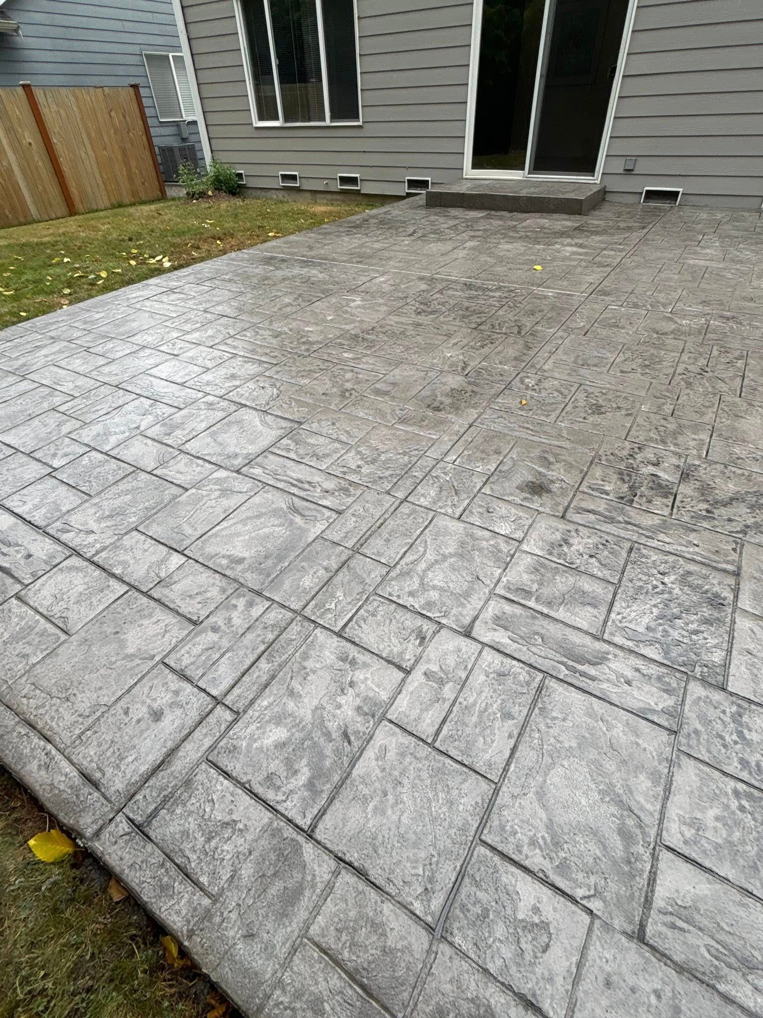Stamped concrete patio in front of a gray house with a sliding glass door. The patio has a stone-like pattern.