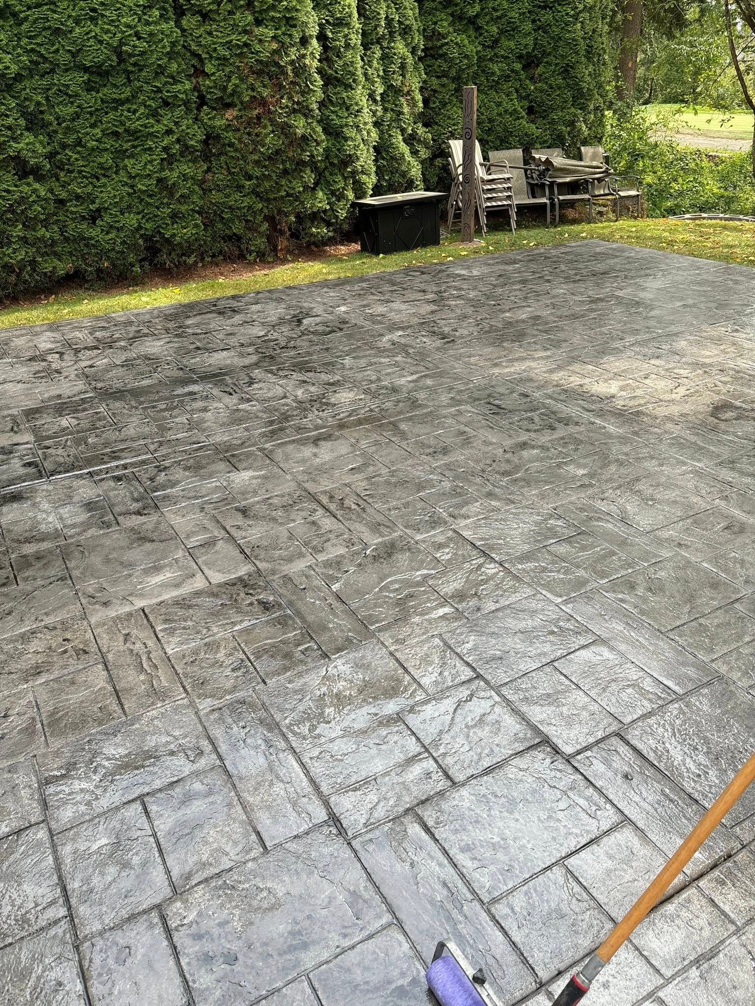 Gray patterned patio with a broom, green bushes in the background.