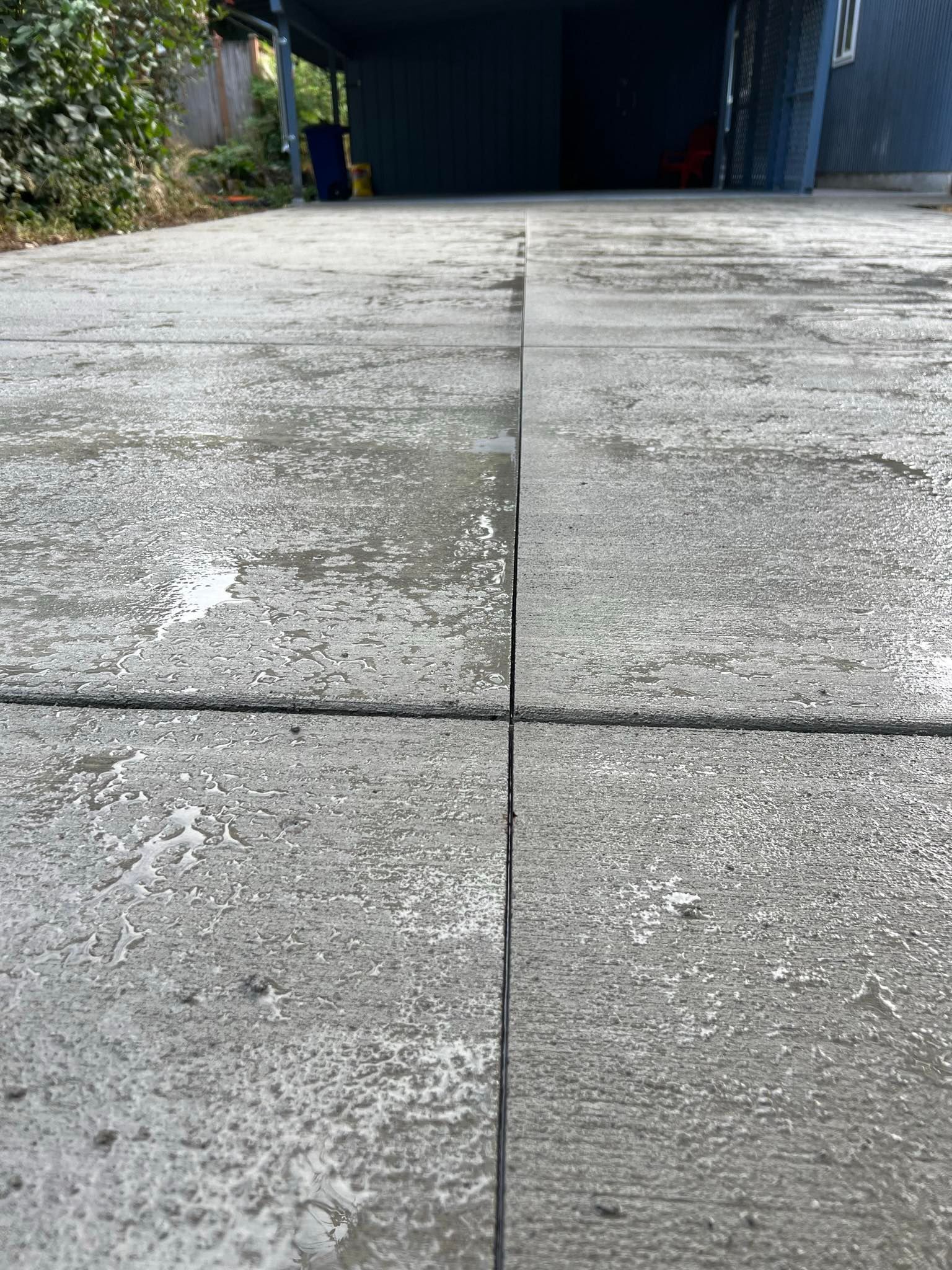 Wet concrete driveway with expansion joints leading towards a covered structure.