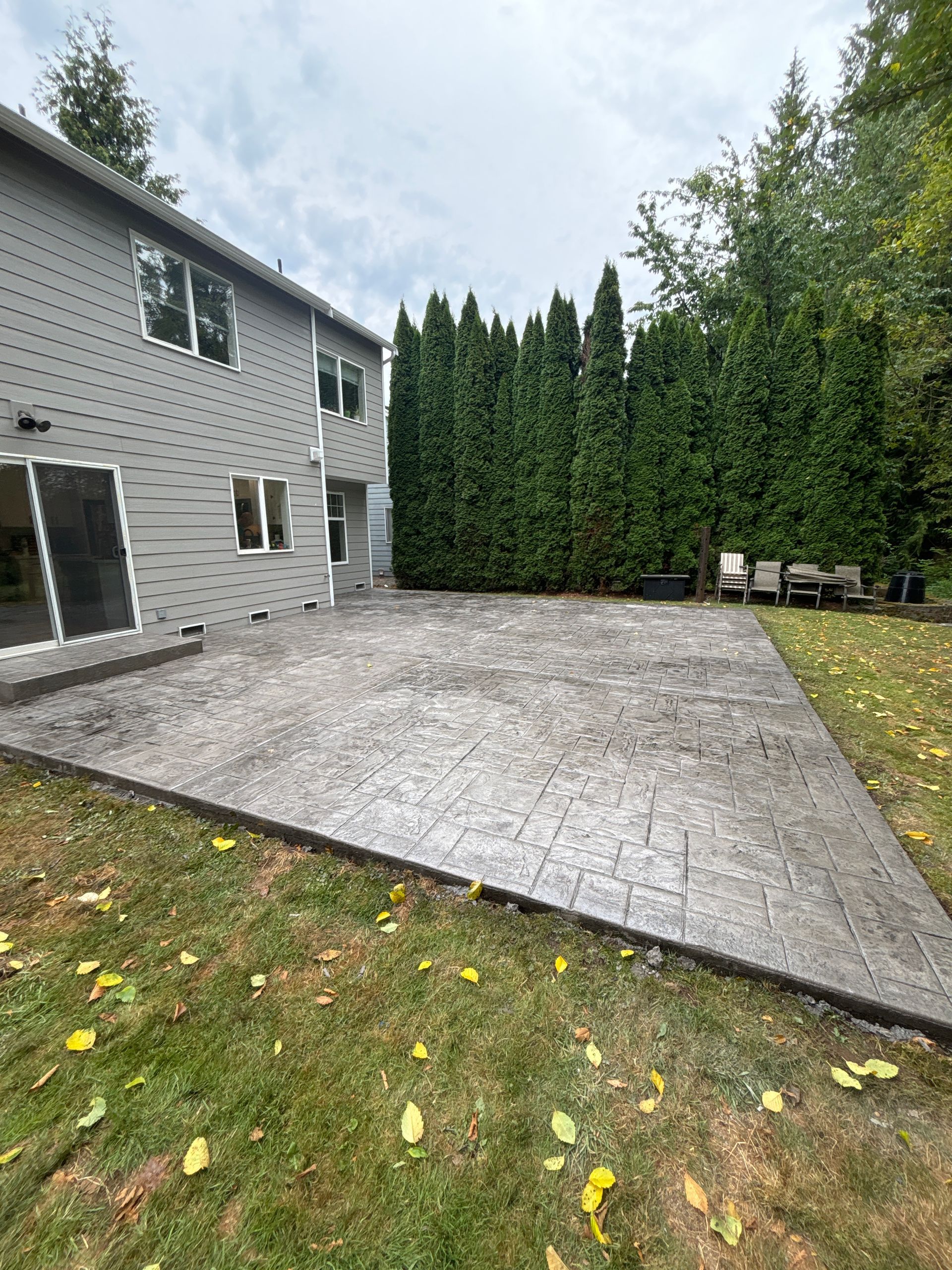 Backyard patio with patterned concrete, bordering a lawn and evergreen hedge. Gray house visible.