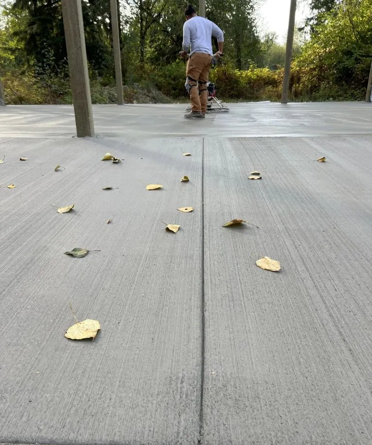 A worker smoothing a fresh concrete surface, with several fallen leaves scattered on the ground.