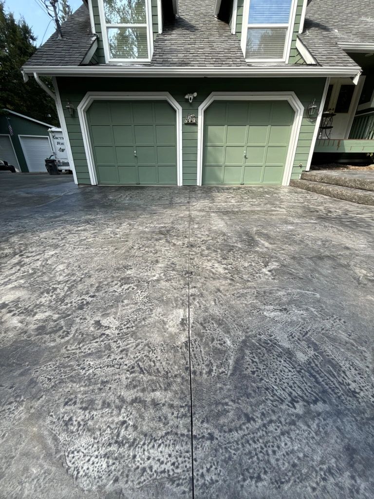 Stamped concrete driveway in front of a house with green garage doors. The driveway has gray mottled patterns and a crack down the middle.