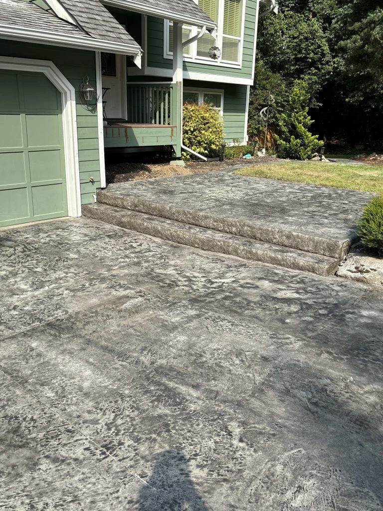 Exterior view of a house with a concrete driveway and steps leading to the entrance. The house is painted green, and the concrete is a mottled gray.