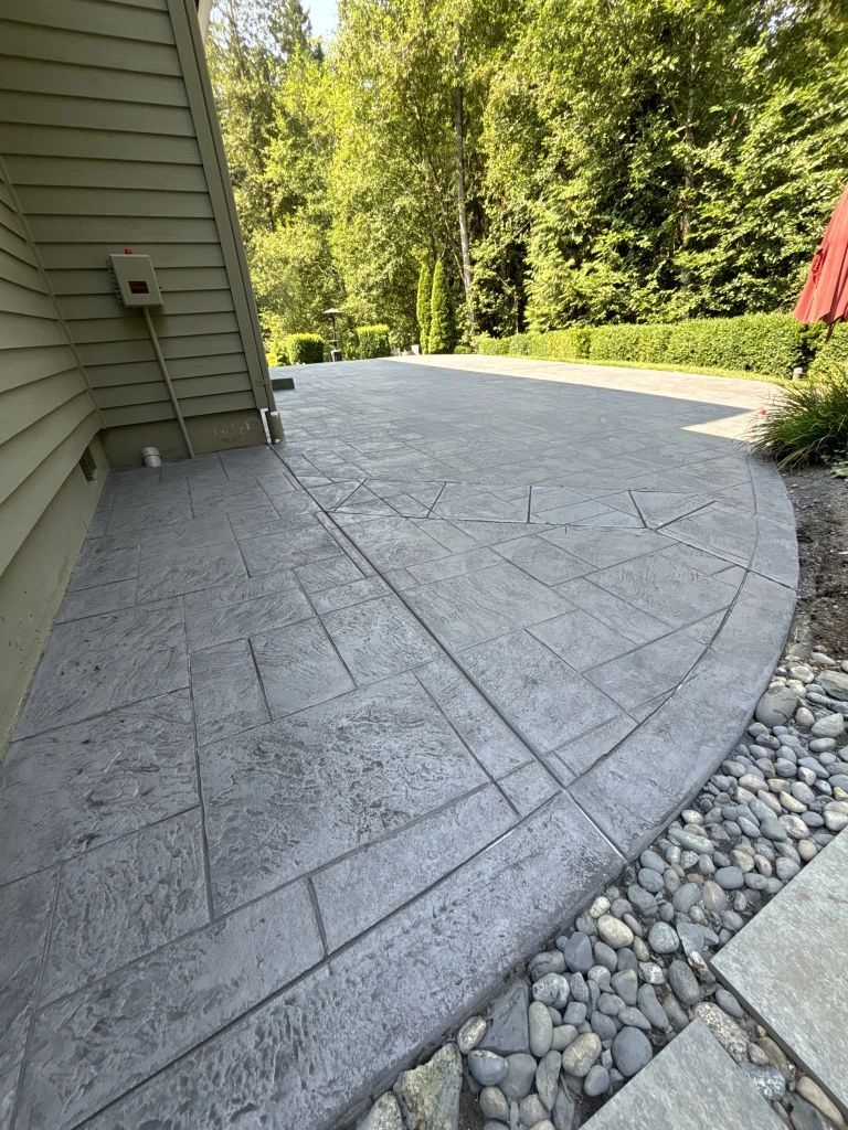 A gray stamped concrete patio curves alongside a house and a gravel border. Green trees and foliage fill the background.